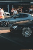 A car meet scene with a gathering of people interacting around high-performance cars. A sleek, silver sports car and a shiny black car with intricate wheel designs are prominently displayed. In the background, several people are standing, some of whom are engaging in conversation or capturing photos. The overall setting suggests a casual, outdoor environment near a building with large windows.