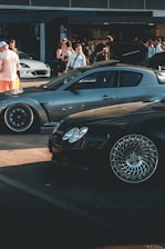 A car meet scene with a gathering of people interacting around high-performance cars. A sleek, silver sports car and a shiny black car with intricate wheel designs are prominently displayed. In the background, several people are standing, some of whom are engaging in conversation or capturing photos. The overall setting suggests a casual, outdoor environment near a building with large windows.