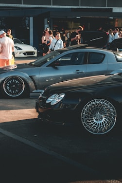 A car meet scene with a gathering of people interacting around high-performance cars. A sleek, silver sports car and a shiny black car with intricate wheel designs are prominently displayed. In the background, several people are standing, some of whom are engaging in conversation or capturing photos. The overall setting suggests a casual, outdoor environment near a building with large windows.