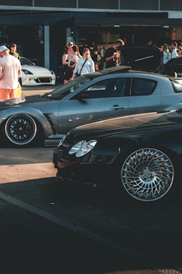 A car meet scene with a gathering of people interacting around high-performance cars. A sleek, silver sports car and a shiny black car with intricate wheel designs are prominently displayed. In the background, several people are standing, some of whom are engaging in conversation or capturing photos. The overall setting suggests a casual, outdoor environment near a building with large windows.