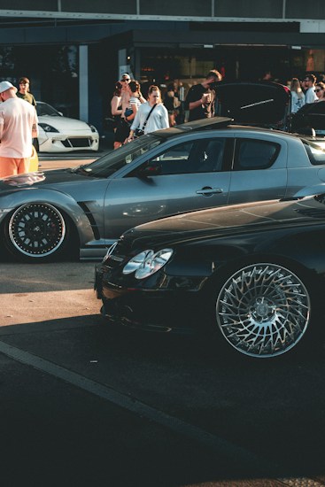 A car meet scene with a gathering of people interacting around high-performance cars. A sleek, silver sports car and a shiny black car with intricate wheel designs are prominently displayed. In the background, several people are standing, some of whom are engaging in conversation or capturing photos. The overall setting suggests a casual, outdoor environment near a building with large windows.
