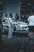 A custom-decorated car is displayed on a platform with graphics and text around it. Several people are gathered around the vehicle, possibly at a car show or exhibition. Confetti is scattered on the ground, adding to the celebratory or festive atmosphere.