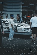 A custom-decorated car is displayed on a platform with graphics and text around it. Several people are gathered around the vehicle, possibly at a car show or exhibition. Confetti is scattered on the ground, adding to the celebratory or festive atmosphere.