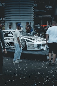 A custom-decorated car is displayed on a platform with graphics and text around it. Several people are gathered around the vehicle, possibly at a car show or exhibition. Confetti is scattered on the ground, adding to the celebratory or festive atmosphere.