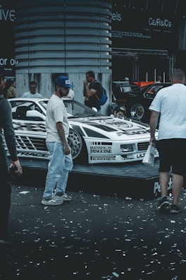 A custom-decorated car is displayed on a platform with graphics and text around it. Several people are gathered around the vehicle, possibly at a car show or exhibition. Confetti is scattered on the ground, adding to the celebratory or festive atmosphere.
