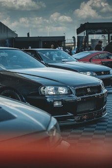 A group of sleek sports cars are parked in an outdoor setting with a cloudy sky overhead. The focus is on a black car with distinctive headlights and a grille emblem. Other cars and people are visible in the background, indicating a car show or gathering.