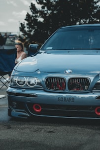 A sleek, customized BMW car with a dark finish, featuring red accents on the front grille and bumper, and the words 'GO HARD' printed on the bumper. A person is seated in the background on a foldable chair beneath a tree, with additional vehicles around, suggesting a car meet or exhibition.