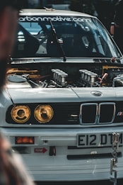 A close-up view of the front of a modified BMW car with its hood open, showcasing the engine and internal components. The car has a white exterior with distinctive headlights and a license plate reading 'E12 DTM'. The windshield displays a website URL decal. The surrounding environment suggests a car event or gathering.