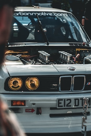 A close-up view of the front of a modified BMW car with its hood open, showcasing the engine and internal components. The car has a white exterior with distinctive headlights and a license plate reading 'E12 DTM'. The windshield displays a website URL decal. The surrounding environment suggests a car event or gathering.
