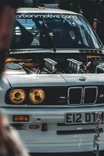 A close-up view of the front of a modified BMW car with its hood open, showcasing the engine and internal components. The car has a white exterior with distinctive headlights and a license plate reading 'E12 DTM'. The windshield displays a website URL decal. The surrounding environment suggests a car event or gathering.