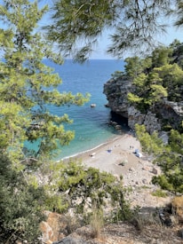 A small, secluded beach surrounded by lush green trees and rocky cliffs. The crystal-clear blue water gently laps at the shore where people are sunbathing and enjoying the water. A few umbrellas and beach towels are spread out on the sand, creating a serene and relaxing atmosphere.