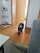 A small, fluffy black and white dog walks across a wooden floor in a cozy home environment. The background features an open doorway leading to another room. On the left, there is a white decorative shelf displaying various items, including small plush toys and candles. The atmosphere appears warm and inviting with natural light entering from the adjacent room.