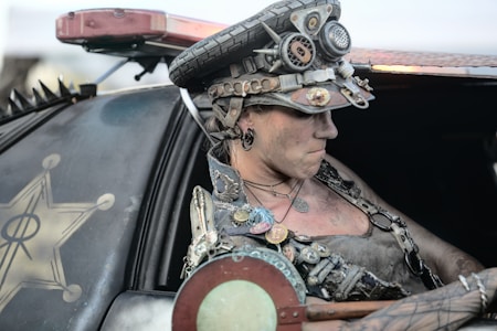A person dressed in a post-apocalyptic or steampunk style is seated in a modified vehicle. The outfit includes a hat adorned with gears and mechanical elements, and the person's clothing features various metallic accessories and patches. The vehicle has a rugged, customized design with visible spikes and a distinctive emblem on its door.