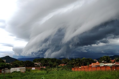 A dramatic storm cloud formation looms over a small town. The sky is filled with thick, dark clouds that swirl in a turbulent pattern. Below, buildings and greenery occupy the landscape, contrasting with the foreboding sky.
