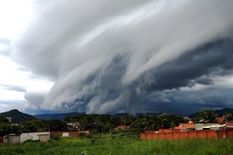 A dramatic storm cloud formation looms over a small town. The sky is filled with thick, dark clouds that swirl in a turbulent pattern. Below, buildings and greenery occupy the landscape, contrasting with the foreboding sky.