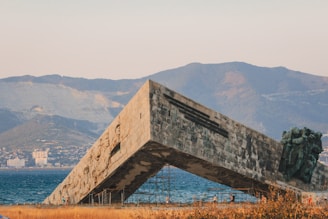A large, angular concrete monument is set against a backdrop of mountains and a body of water. The structure features sculpted human figures along one side and stands tilted towards the ground. A few people are walking nearby, providing a sense of scale. The surroundings include sparse vegetation, and the scene is captured in warm, late-afternoon lighting.