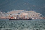 A large ship is anchored in a body of water with a busy industrial facility in the background. The facility includes numerous structures, machinery, and cranes, set against a backdrop of forested mountains.