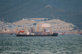 A large ship is anchored in a body of water with a busy industrial facility in the background. The facility includes numerous structures, machinery, and cranes, set against a backdrop of forested mountains.