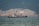 A large ship is anchored in a body of water with a busy industrial facility in the background. The facility includes numerous structures, machinery, and cranes, set against a backdrop of forested mountains.
