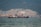 A large ship is anchored in a body of water with a busy industrial facility in the background. The facility includes numerous structures, machinery, and cranes, set against a backdrop of forested mountains.