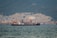 A large ship is anchored in a body of water with a busy industrial facility in the background. The facility includes numerous structures, machinery, and cranes, set against a backdrop of forested mountains.