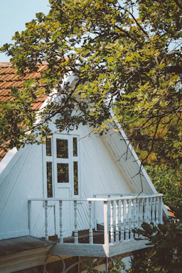 A quaint white A-frame house with a small balcony surrounded by leafy green tree branches. The house has a rustic, wooden facade with a sloped roof. Sunlight filters through the leaves, casting gentle shadows on the building.