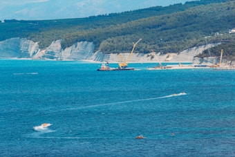 A coastal scene features a large construction barge equipped with a crane near a rugged shoreline. The turquoise sea dominates the foreground, with several small boats visible, including a speedboat leaving a white wake. In the background, steep cliffs and forested hills form the horizon under a clear sky.