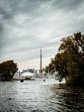 A body of water with the CN Tower prominently visible against a cloudy sky. The skyline includes modern buildings in the background, with dense trees on either side of the photograph framing the scene.