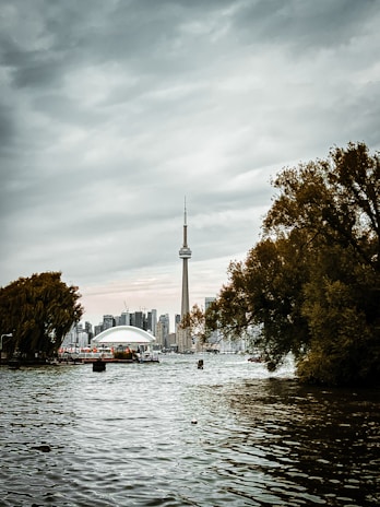 A body of water with the CN Tower prominently visible against a cloudy sky. The skyline includes modern buildings in the background, with dense trees on either side of the photograph framing the scene.