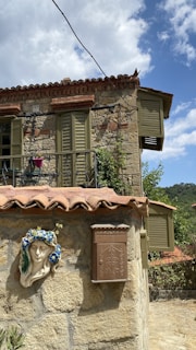 A rustic stone house with olive green shutters and terracotta roof tiles is seen. The facade features a decorative relief of a face adorned with a flower crown. A brown mailbox with decorative detailing is mounted on the wall. The background shows lush greenery and a clear blue sky with clouds.