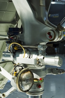 Close-up view of a metal mechanical component, likely part of an aircraft's landing gear assembly, featuring various wires, bolts, and a round light fixture. The image is filled with metallic textures and a mix of smooth and structured surfaces.