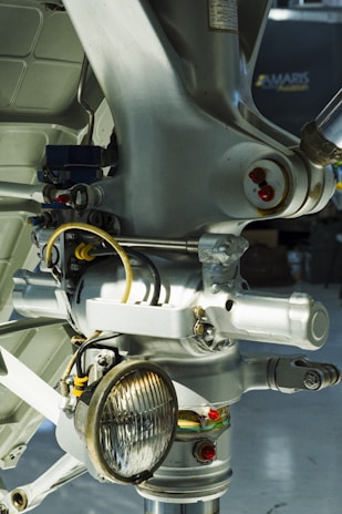 Close-up view of a metal mechanical component, likely part of an aircraft's landing gear assembly, featuring various wires, bolts, and a round light fixture. The image is filled with metallic textures and a mix of smooth and structured surfaces.