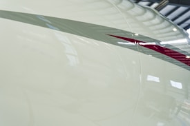 A close-up view of an aircraft's exterior with a smooth, glossy surface. A sleek, maroon and gray stripe runs diagonally across the cream-colored body. The background reveals the metallic structure of a hangar, with reflections of light visible on the plane's surface.
