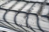 A close-up view of a reflective, textured surface with evenly spaced circular indentations. Light and shadow create an intricate pattern formed by the reflection of a grid or framework above. The surface has a smooth metallic appearance, with subtle reflections adding depth.