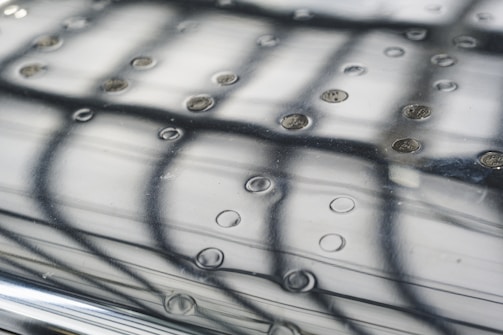 A close-up view of a reflective, textured surface with evenly spaced circular indentations. Light and shadow create an intricate pattern formed by the reflection of a grid or framework above. The surface has a smooth metallic appearance, with subtle reflections adding depth.