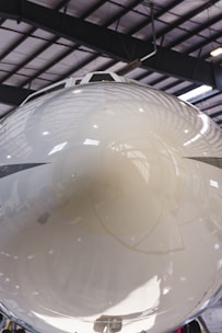 A close-up view of the reflective nose cone of an aircraft inside a hangar. The polished surface shows reflections of the interior, including structural beams and lighting. The setting appears industrial with a large, spacious ceiling.