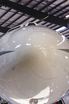A close-up view of the reflective nose cone of an aircraft inside a hangar. The polished surface shows reflections of the interior, including structural beams and lighting. The setting appears industrial with a large, spacious ceiling.