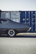The rear section of a classic, dark-colored car is parked near large blue and gray shipping containers against a clear sky. The vehicle's shiny wheel with radial lettering is prominently visible, contrasting with the rugged industrial setting.