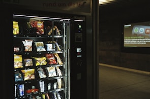 A vending machine is stocked with various snack items, predominantly chips and candy. There is a digital display to the right, suggesting it's located in a public space like a subway station, with an advertisement displayed on the wall.