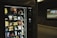 A vending machine is stocked with various snack items, predominantly chips and candy. There is a digital display to the right, suggesting it's located in a public space like a subway station, with an advertisement displayed on the wall.