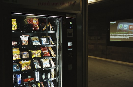 A vending machine is stocked with various snack items, predominantly chips and candy. There is a digital display to the right, suggesting it's located in a public space like a subway station, with an advertisement displayed on the wall.