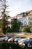 Several boats covered with tarps are moored along a calm waterway. The background features a charming blue building adorned with shutters, surrounded by lush greenery. The scene is bathed in warm sunlight, creating a serene and picturesque setting.