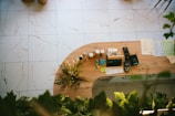 An overhead view of a wooden reception desk with neatly arranged items including bottles, plants, a register, and several sheets of paper. The floor is tiled with a marble-like pattern, and there is greenery partially visible in the foreground.