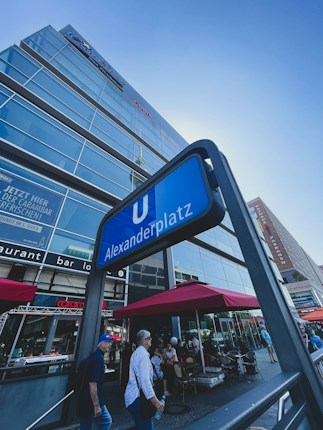 An urban scene featuring a glass-fronted building rising towards the sky, with reflections visible on its surface. In the foreground, a sign indicates Alexanderplatz, a well-known location. People walk along the street and sit at an outdoor cafe under red umbrellas.