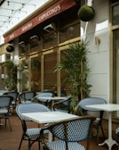 An outdoor café area with blue and white checkered chairs and white square tables. The café has a red awning with signs displaying 'Pastas', 'Fresh Fish', and 'Cappuccino's'. Green plants are placed along the walls and string lights are hung above.