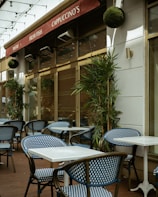 An outdoor café area with blue and white checkered chairs and white square tables. The café has a red awning with signs displaying 'Pastas', 'Fresh Fish', and 'Cappuccino's'. Green plants are placed along the walls and string lights are hung above.