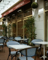 An outdoor café area with blue and white checkered chairs and white square tables. The café has a red awning with signs displaying 'Pastas', 'Fresh Fish', and 'Cappuccino's'. Green plants are placed along the walls and string lights are hung above.