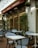 An outdoor café area with blue and white checkered chairs and white square tables. The café has a red awning with signs displaying 'Pastas', 'Fresh Fish', and 'Cappuccino's'. Green plants are placed along the walls and string lights are hung above.