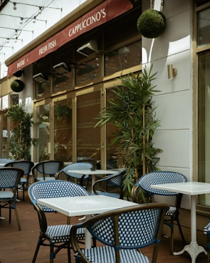 An outdoor café area with blue and white checkered chairs and white square tables. The café has a red awning with signs displaying 'Pastas', 'Fresh Fish', and 'Cappuccino's'. Green plants are placed along the walls and string lights are hung above.