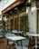 An outdoor café area with blue and white checkered chairs and white square tables. The café has a red awning with signs displaying 'Pastas', 'Fresh Fish', and 'Cappuccino's'. Green plants are placed along the walls and string lights are hung above.